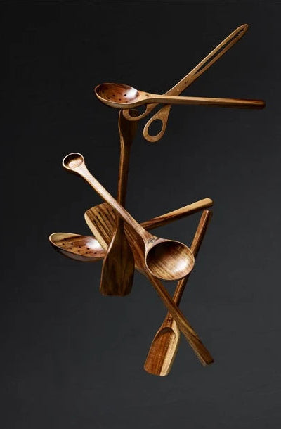 A sculptural wooden spoon rack made of multiple crossed and hanging wooden spoons with visible wood grain, displayed against a dark charcoal background with a light marble surface at the bottom.