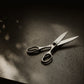 A pair of silver metal scissors with black handles, resting on a dark textured surface under dramatic, directional light that casts a soft shadow.