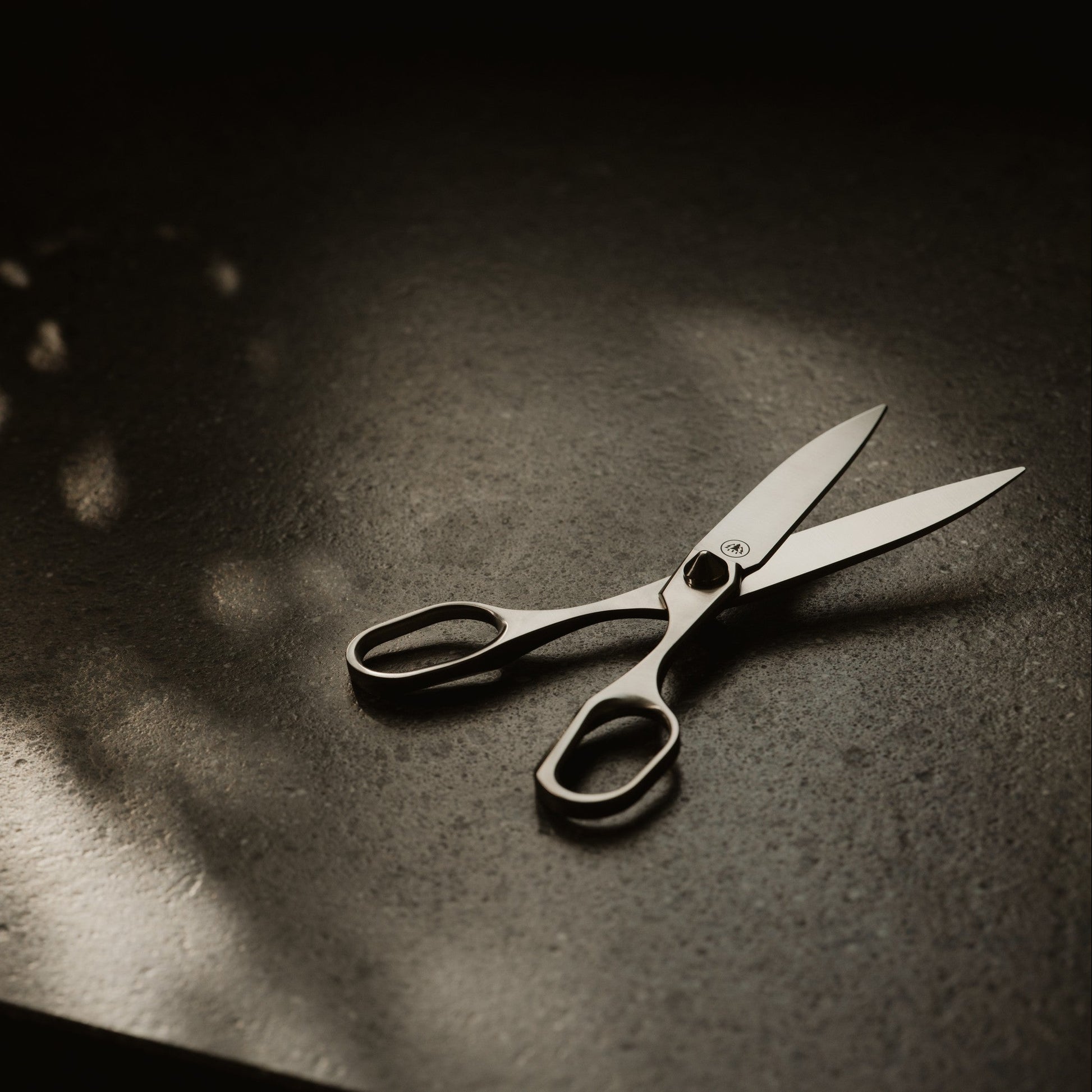 A pair of silver metal scissors with black handles, resting on a dark textured surface under dramatic, directional light that casts a soft shadow.
