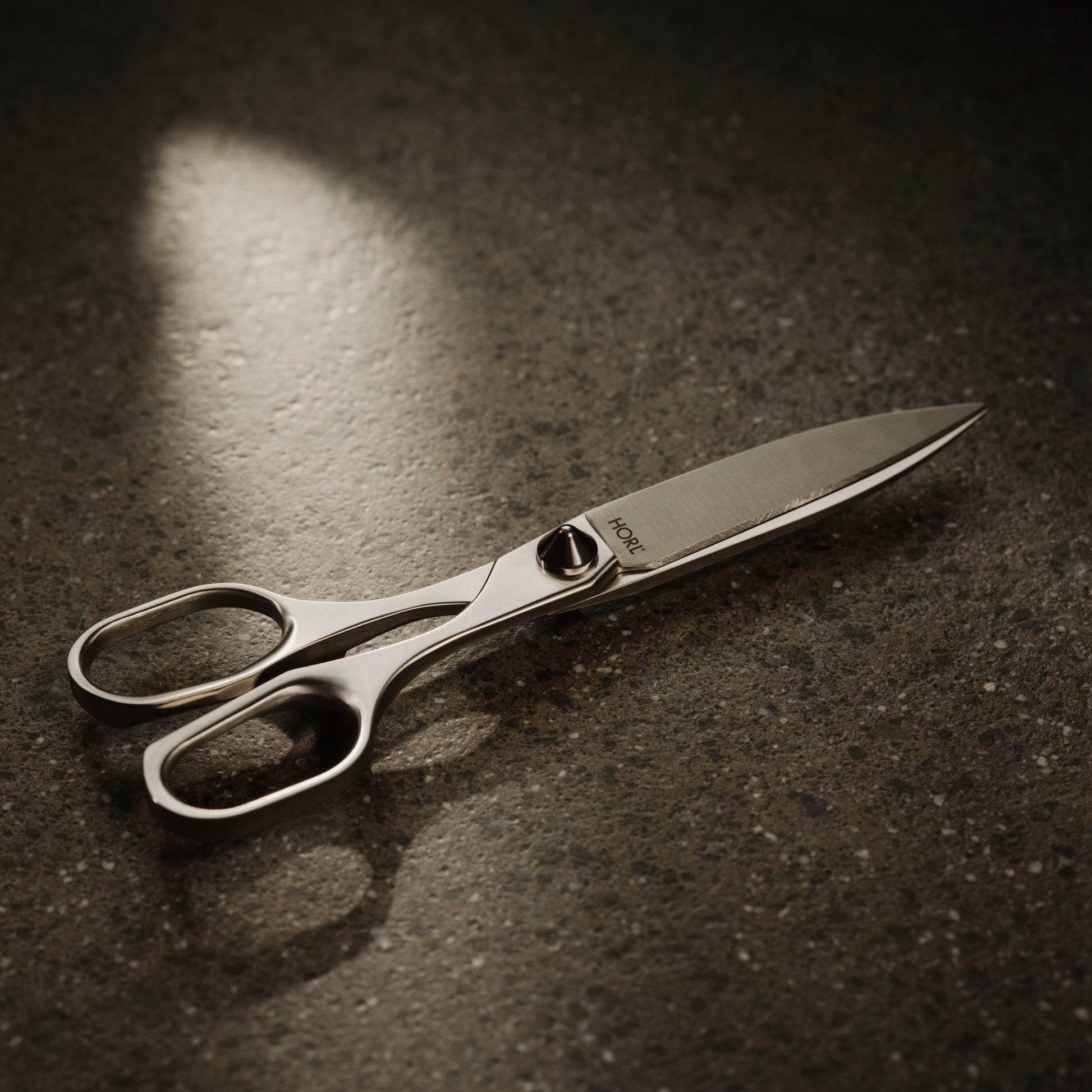 A pair of polished stainless steel scissors with ergonomic finger loops, resting on a dark, textured surface under dramatic spotlighting.