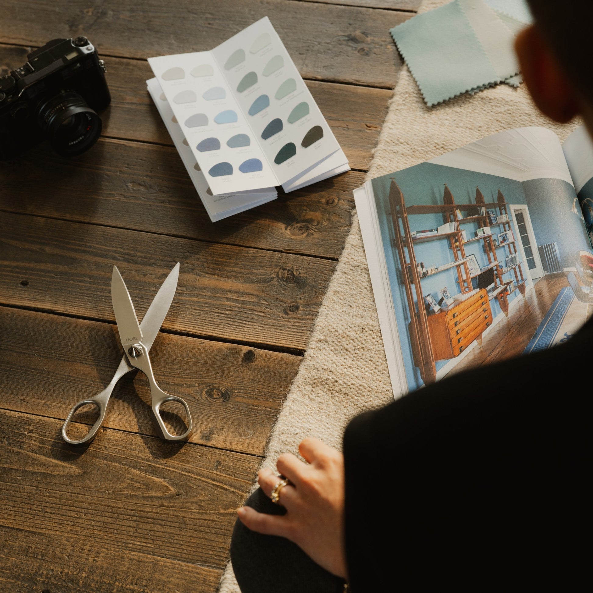 A person viewed from behind sits at a wooden table, holding a swatch book. The table is styled with a camera, scissors, a color swatch book, and an open magazine showing interior design photos.