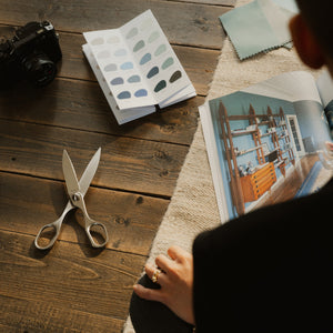 A person viewed from behind sits at a wooden table, holding a swatch book. The table is styled with a camera, scissors, a color swatch book, and an open magazine showing interior design photos.