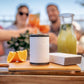 A white reusable travel mug with a black lid sits on a wooden cutting board on an outdoor table, alongside an orange slice, a small white box, and a glass pitcher of juice. Three blurred people are visible in the background toasting.