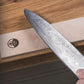 A close-up of a hand holding a Damascus steel chef's knife over a wooden surface with a beige Honkai brand leather sheath.