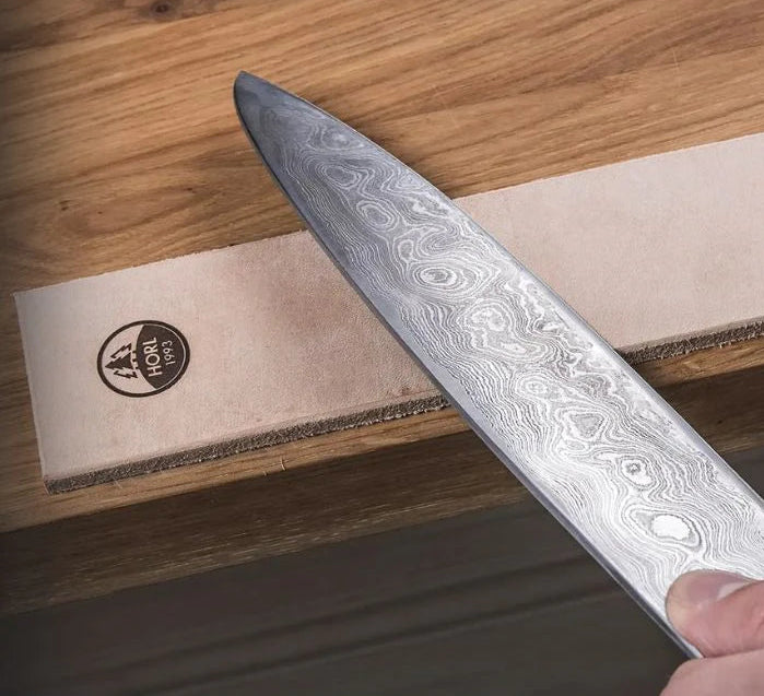 A close-up of a hand holding a Damascus steel chef's knife over a wooden surface with a beige Honkai brand leather sheath.