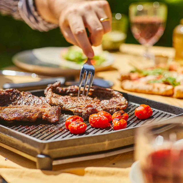 Grilled meat with cherry tomatoes on a grill pan, surrounded by outdoor dining setting.