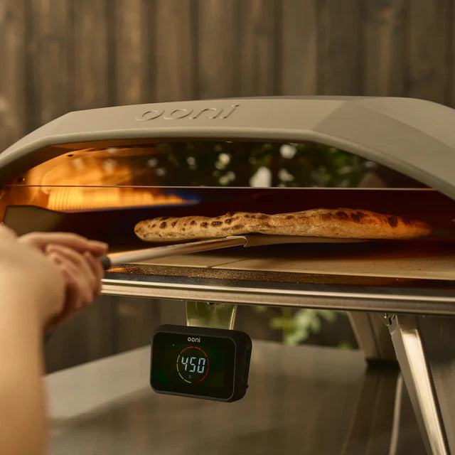 Person taking a pizza out of an open outdoor pizza oven with a digital display showing 450 degrees.