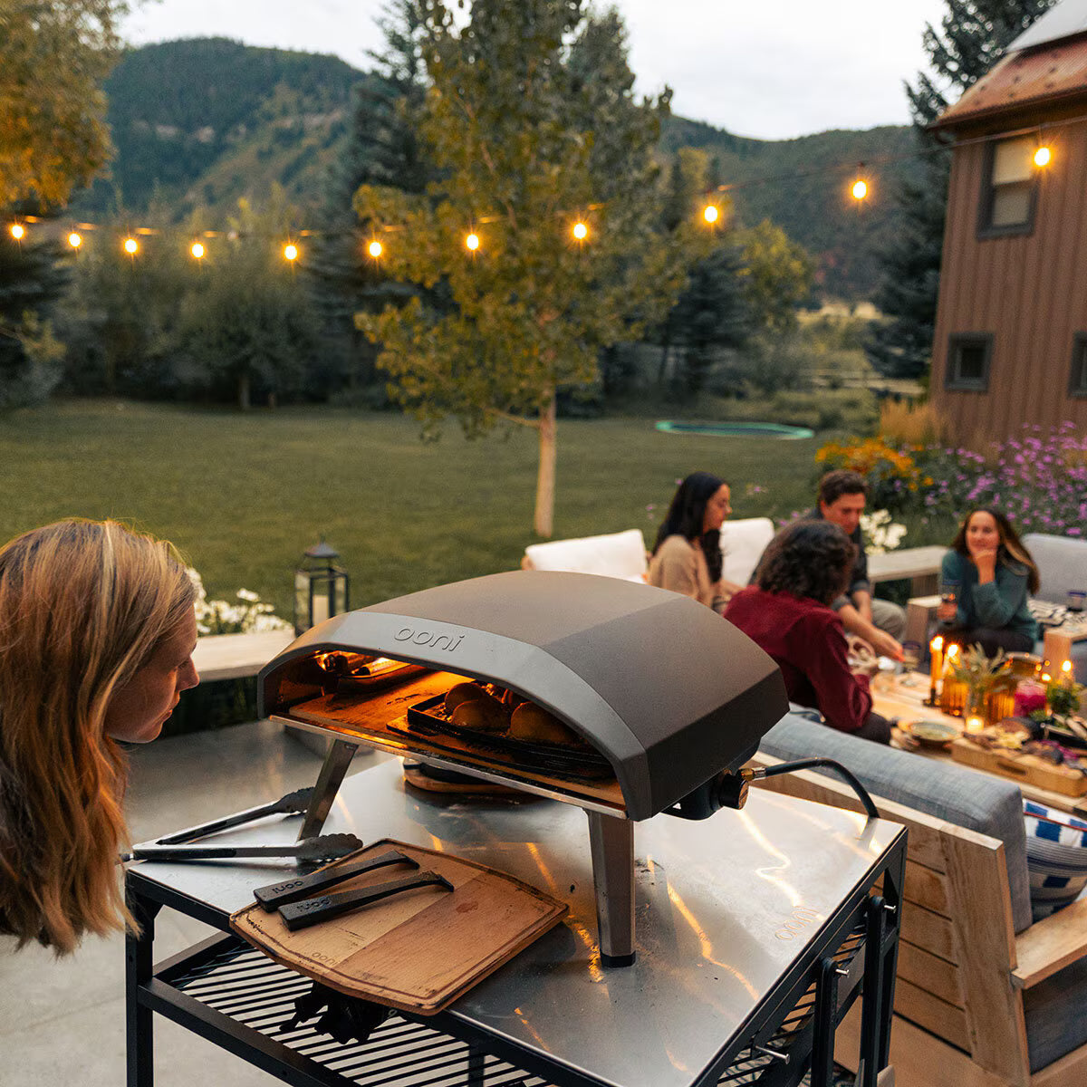 Outdoor setting with a portable pizza oven on a table, people sitting around, and string lights in the background.