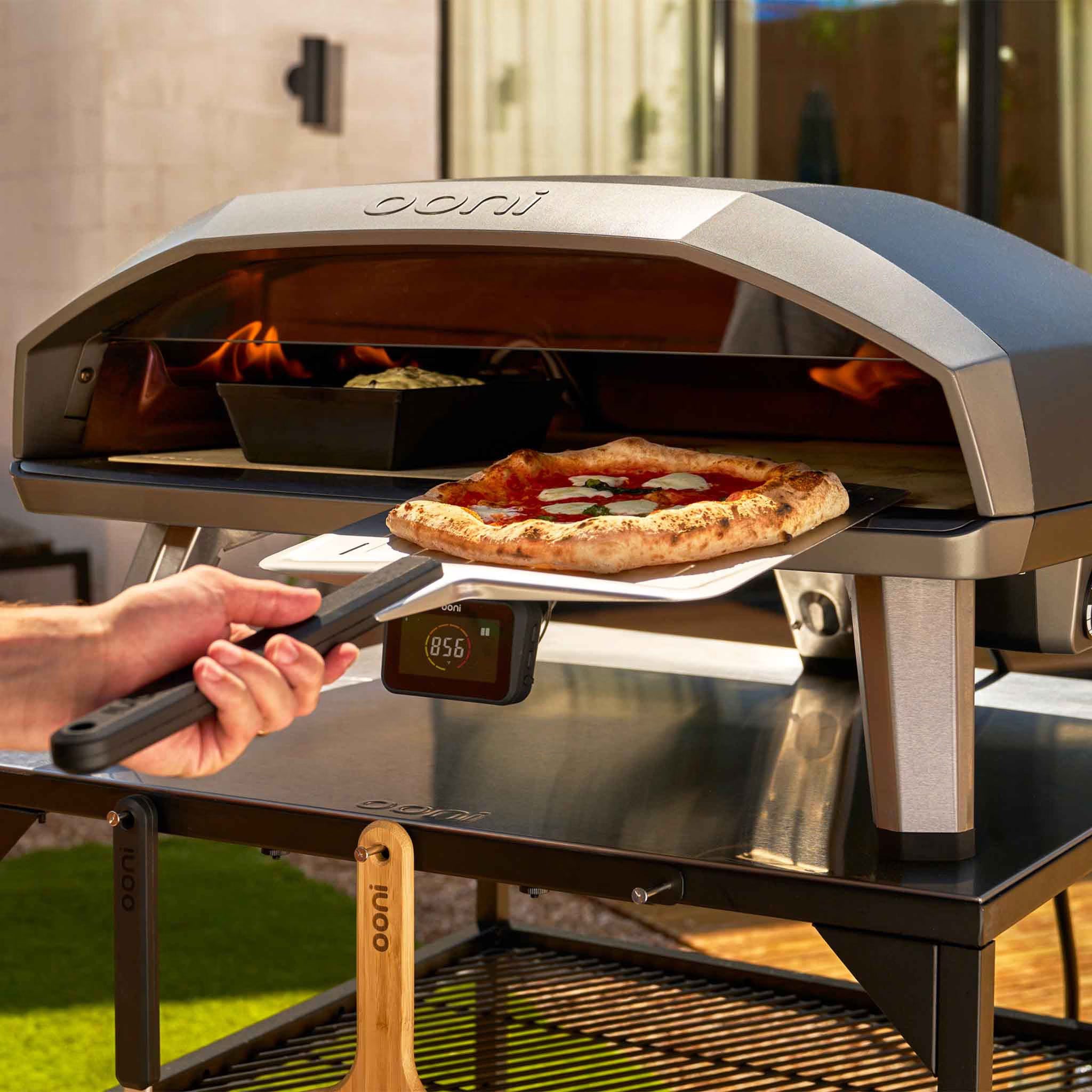 Person placing a pizza into an Ooni pizza oven outdoors.