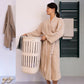 A woman wearing a beige textured bathrobe stands in a bathroom holding a white laundry basket. She is wearing metallic gold slide sandals, with a brown towel hanging on a wall hook to her left and a dark green heated towel rail on the tiled wall to her right.