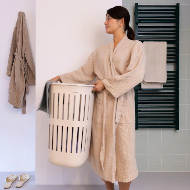 A woman wearing a beige textured bathrobe stands in a bathroom holding a white laundry basket. She is wearing metallic gold slide sandals, with a brown towel hanging on a wall hook to her left and a dark green heated towel rail on the tiled wall to her right.