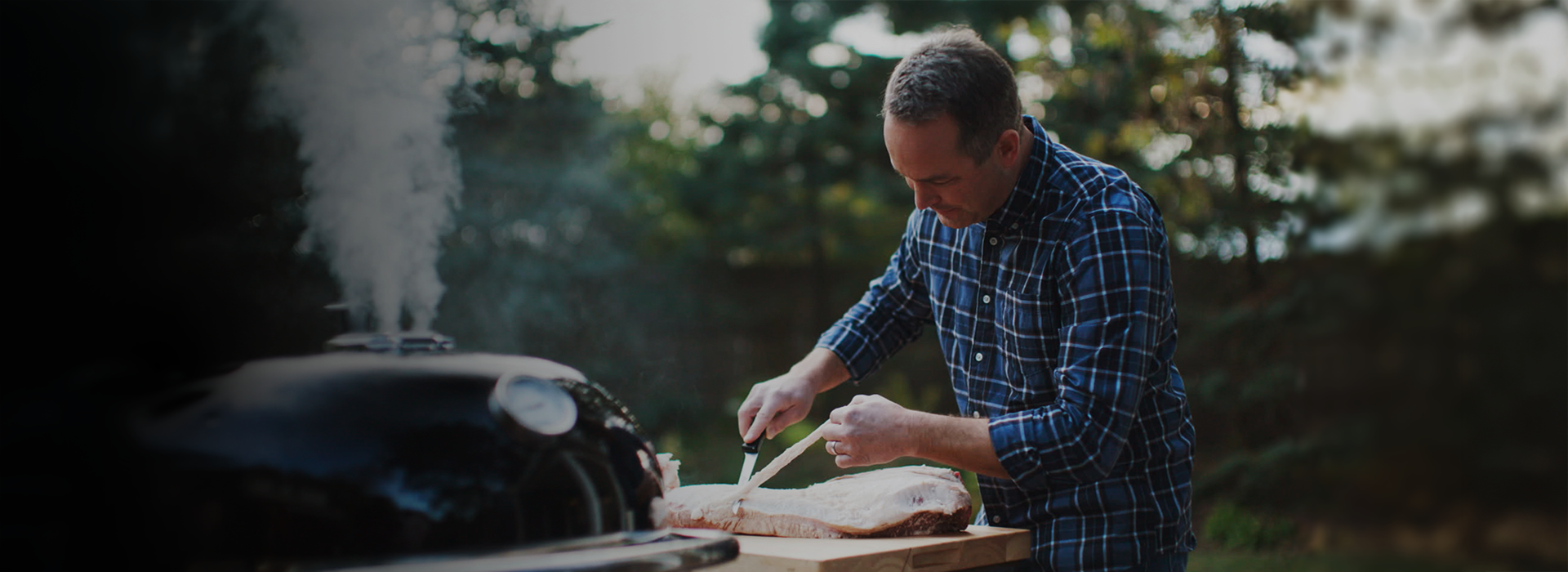 Man preparing food on a grill outdoors with trees in the background