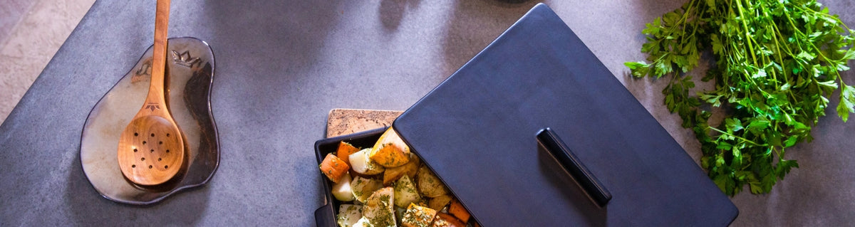 A black cast iron Dutch oven with its lid off, revealing cooked pasta and vegetables inside, styled on a dark gray countertop next to a wooden spoon, fresh parsley, and a dark slate dish.